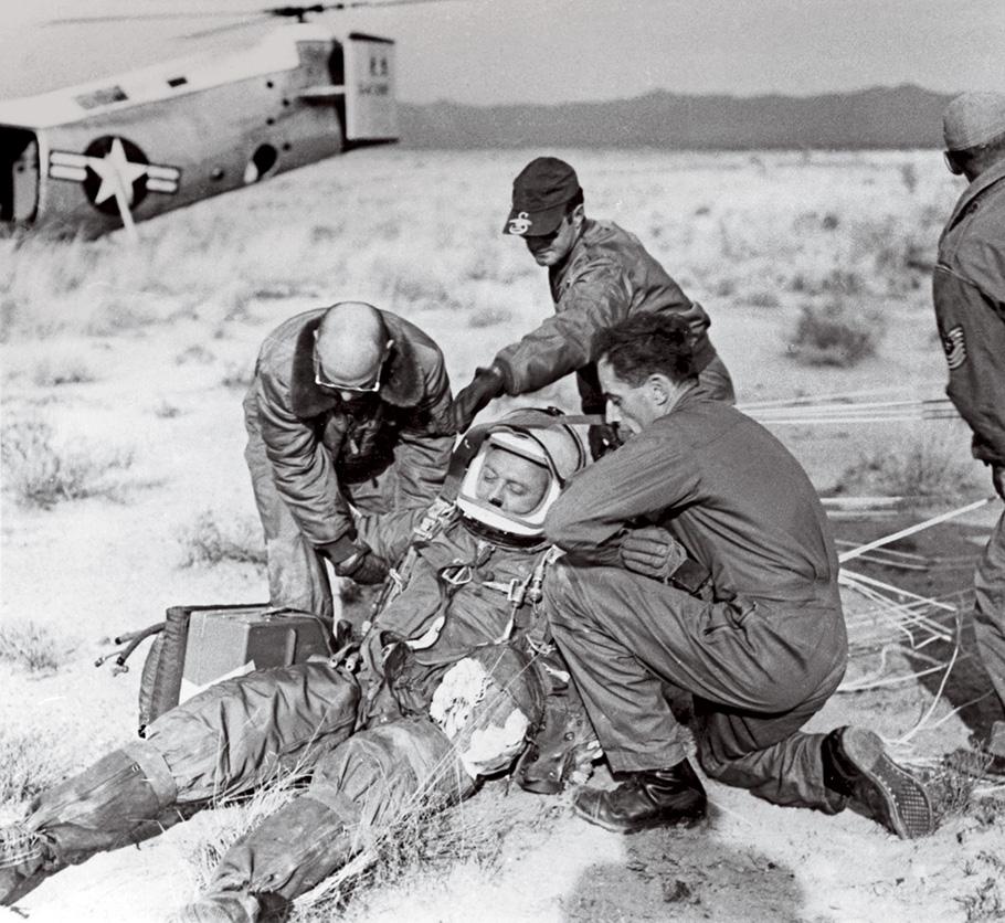 A black and white photograph of a ground crew assisting Capt. Kittinger, a man in a helmet, flight suit, and wearing a parachute. The ground crew helps him remove his flight gear.