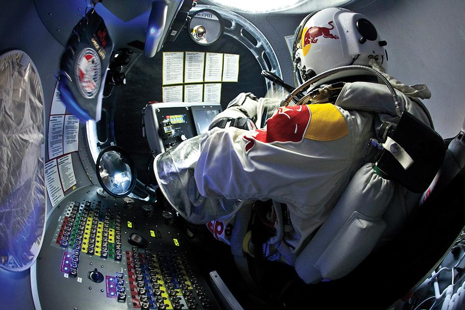 A pilot in a helmet with a red bull on it and a flight, sits inside a small gondola. To their left is a panel of switches. They're preparing to step out from the capsule. 