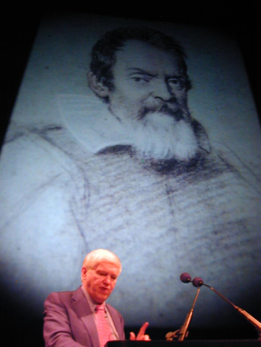 A man speaking into a microphone at the bottom of the frame with a screen of a portrait of a man in the background that makes up the bulk of the image.