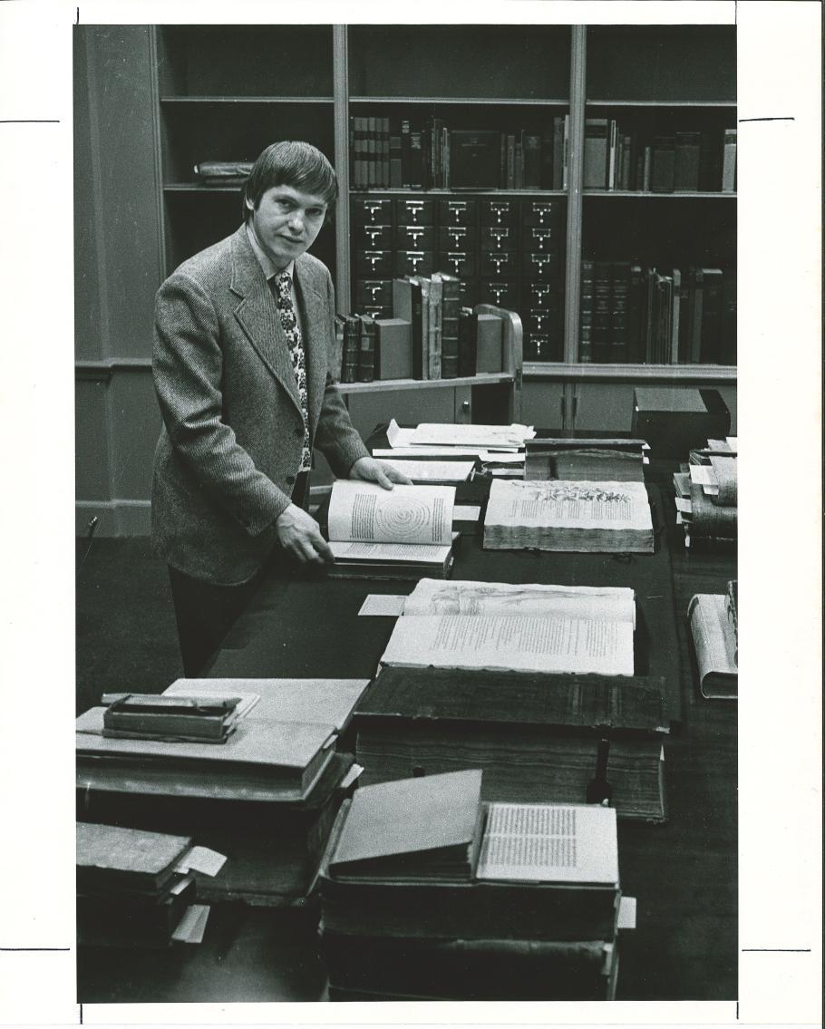 Black and white photo of a man standing by a table with many large books on it.