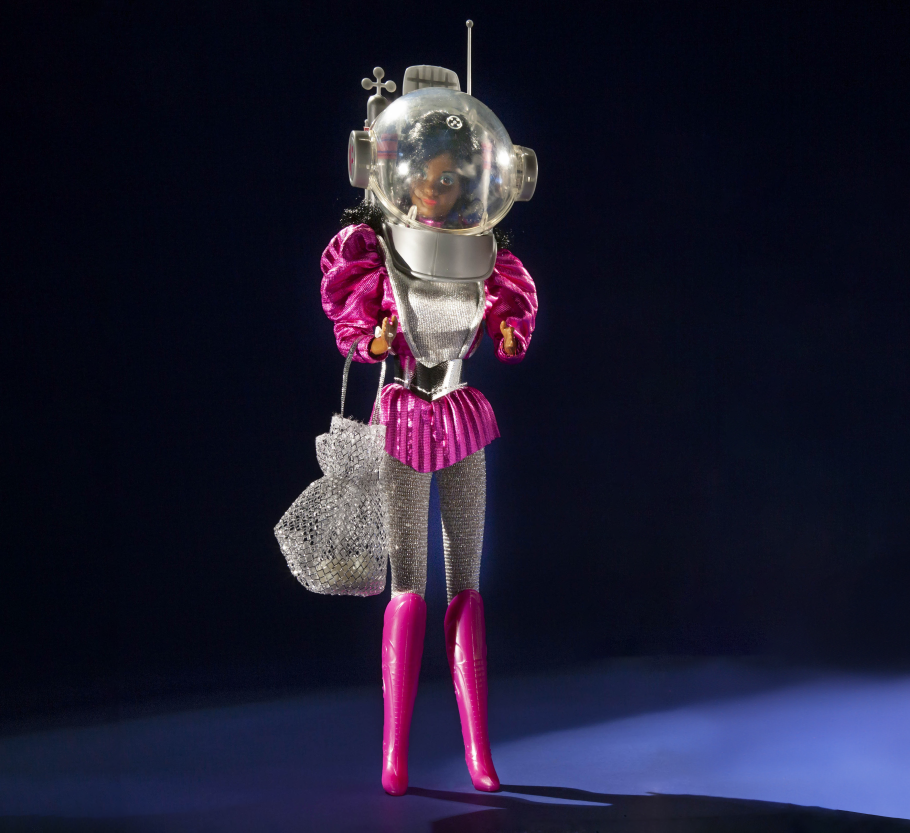 1985 Astronaut Barbie photographed against a dark background. She wears pink boots, silver leggings, a poofy pink top, and a space helmet.