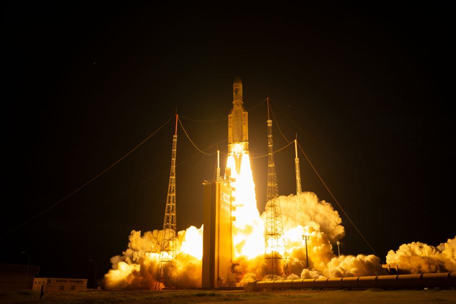 Ariane 5 rocket launches at night. The sky is dark but the area around the rocket is illuminated by the flames from the rocket's engines. Smoke billows around it.
