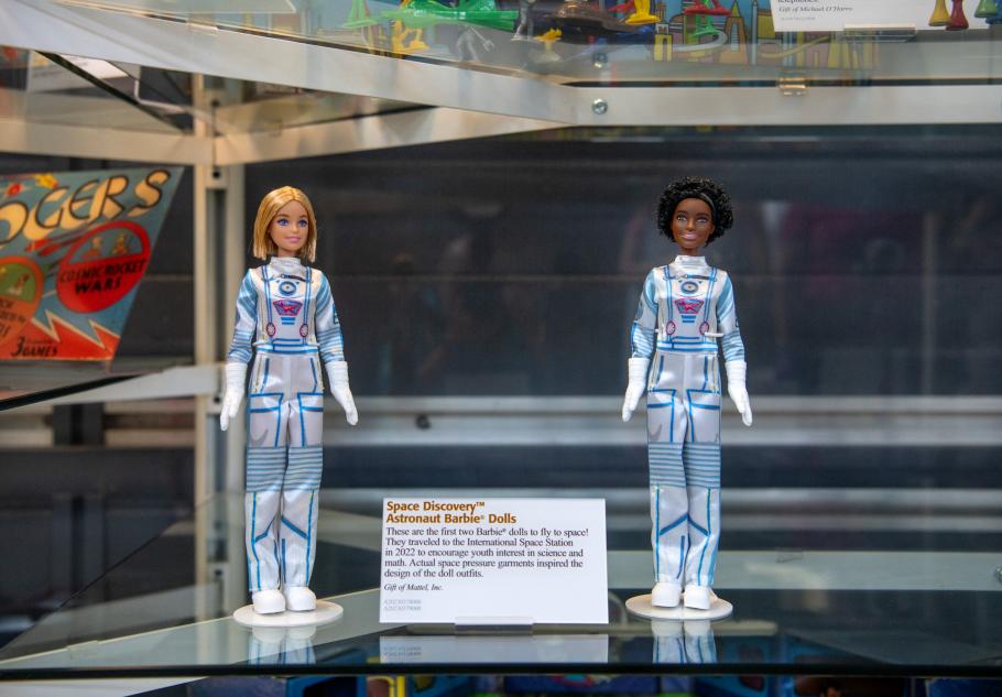 Two Space Discovery Barbies photographed in a glass case. They appear to be standing up straight. Between them is a small artifact description card.