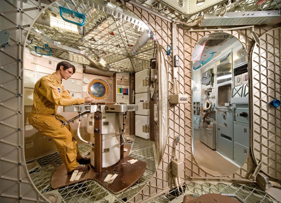 A mannequin sits at a work table inside the backup version of the Skylab orbital workshop.