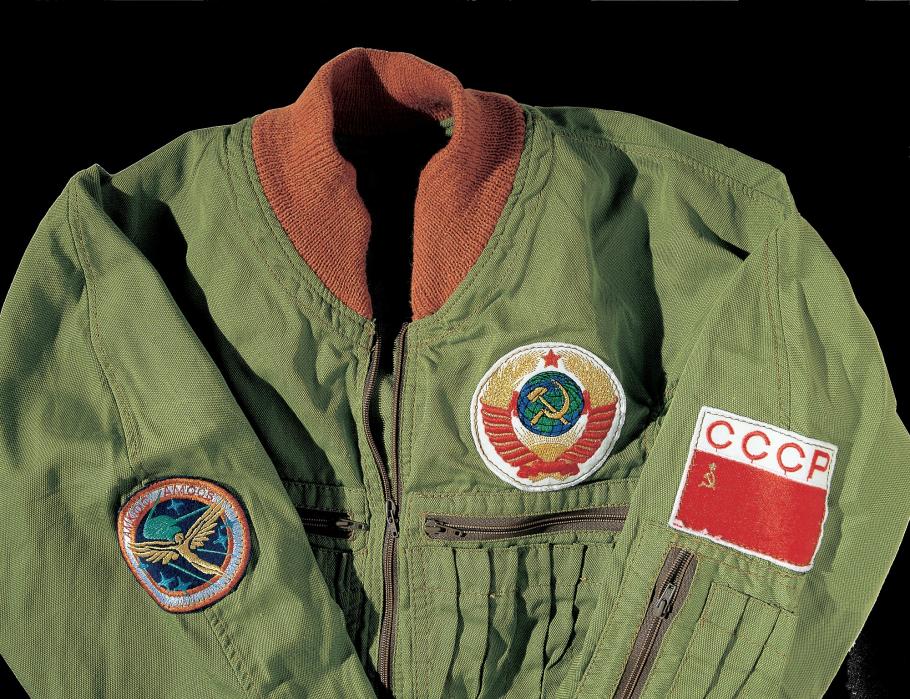 The top half of a green synthetic, woven cloth coverall with burnt orange collar.  The suit has a Soviet flag patch on the left shoulder with the letters CCCP along the top; a patch of the Soviet seal above the left breast pocket; and on the right shoulder is a Salyut space station program patch.