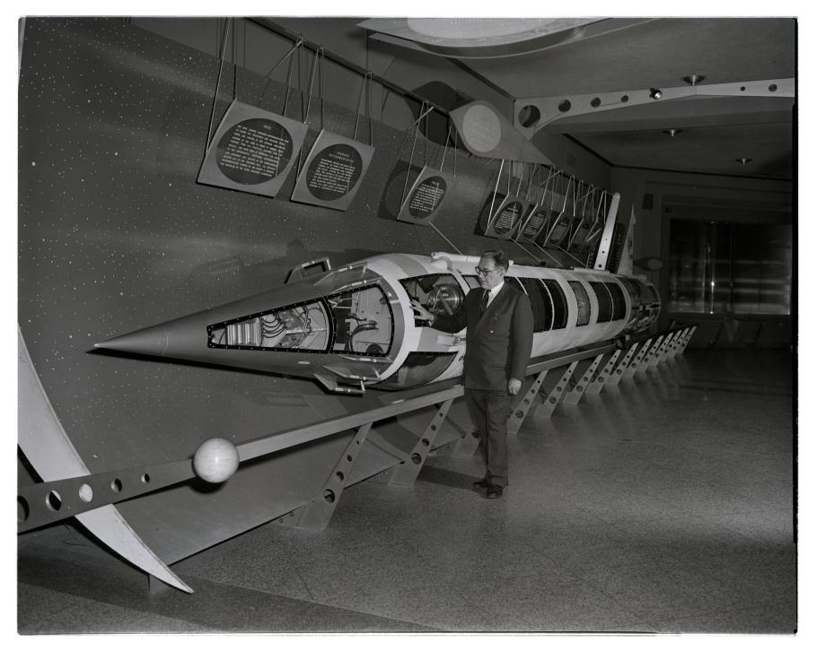 A man stands with one hand on a rocket laying horizontally in a hallway.
