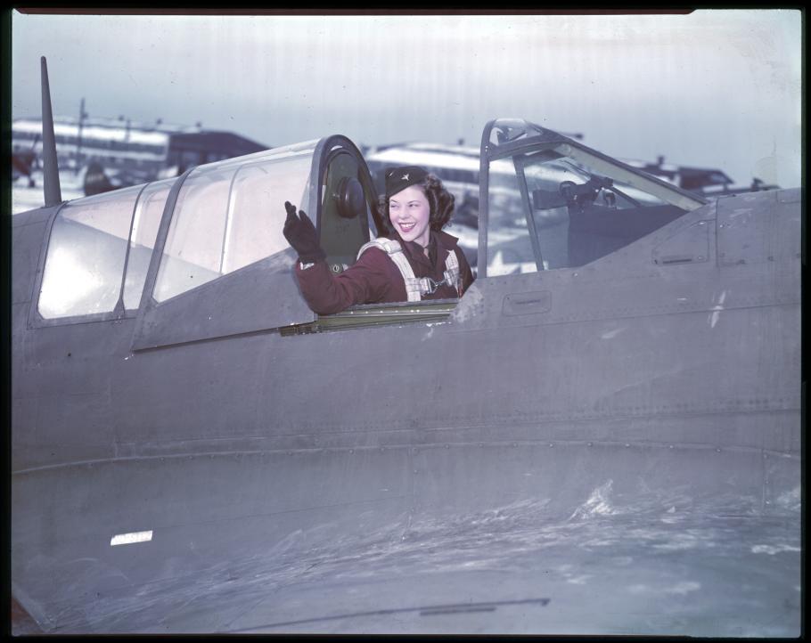 A woman in a military uniform with 1940s styled hair waves from the cockpit of an airplane?