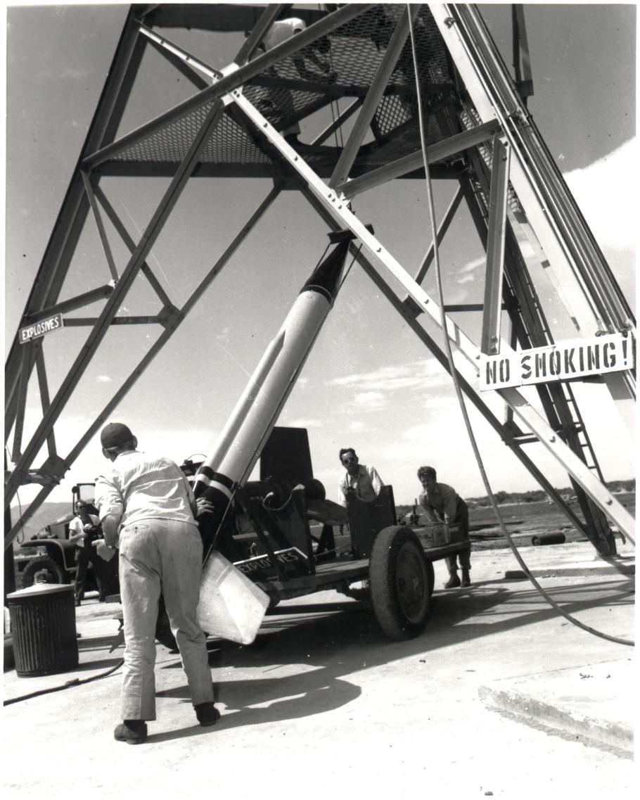 A group of men push a wheeled vehicle underneath a large tower. A rocket sits atop the wheeled vehicle. A sign that says "no smoking!" is attached to the tower.