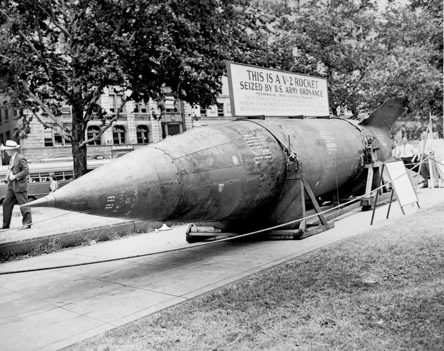 A large rocket lays horizontally on a city street with ropes around it. A sign on top of it states that it is a captured German V-2 rocket.