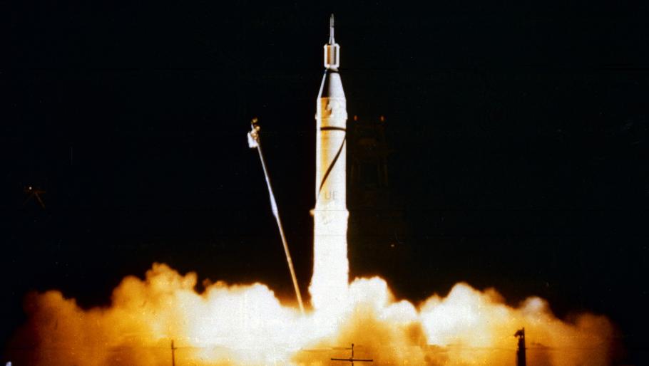 A large rocket taking off of a landing pad with a satellite attached to the top of it. Smoke and exhaust bellow out from the bottom of the rocket.