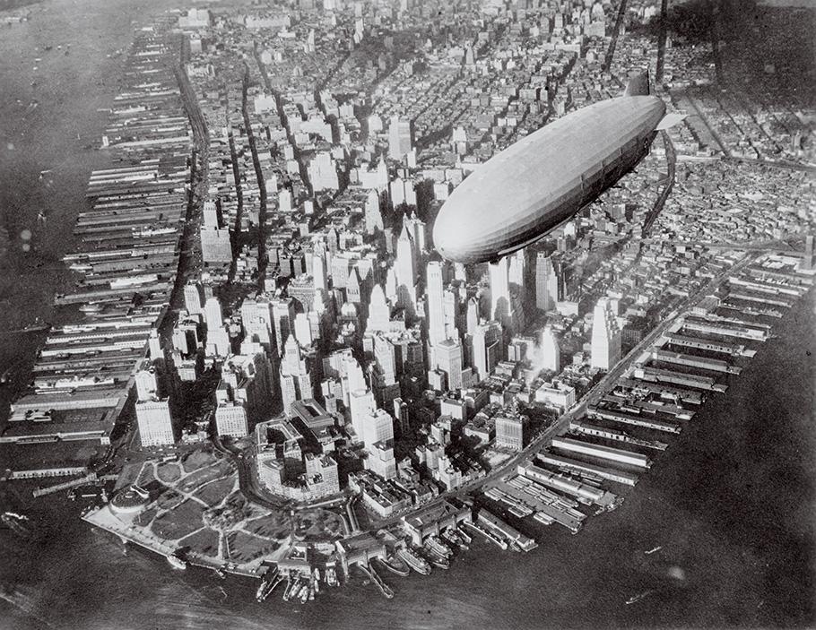 A black-and-white photo shows downtown Manhattan skyscrapers from above, will a white airship edging into the image.