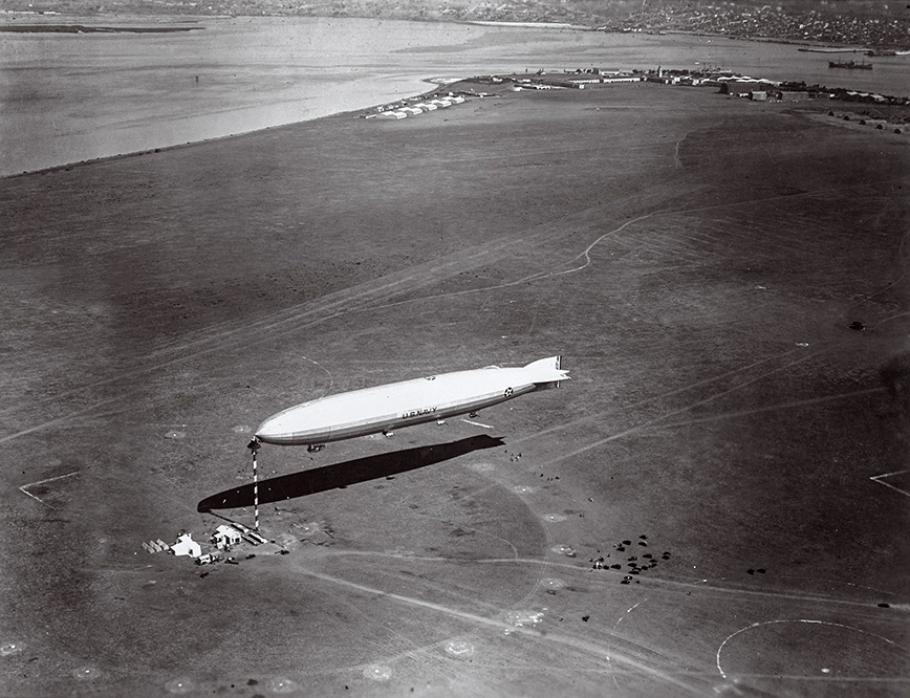 In a black-and-white photo, a white airship casts a dark shadow on the ground. The surrounding landscape is mostly barren except for some buildings in the distance.