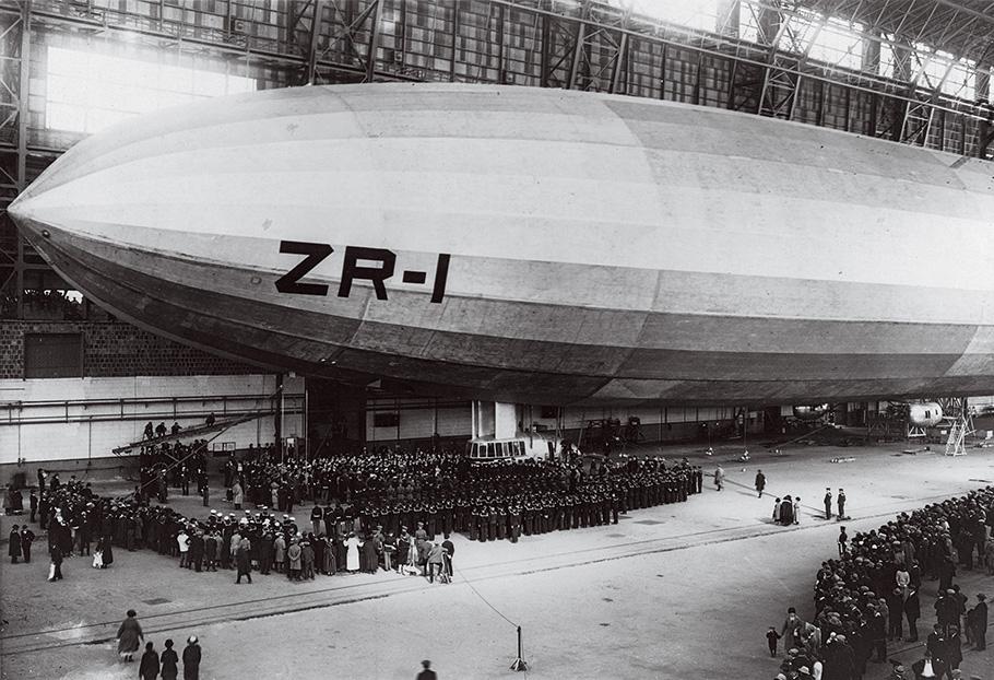 The black and white photo shows a crowd of people dwarfed by a massive airship with the marking "ZR-1"