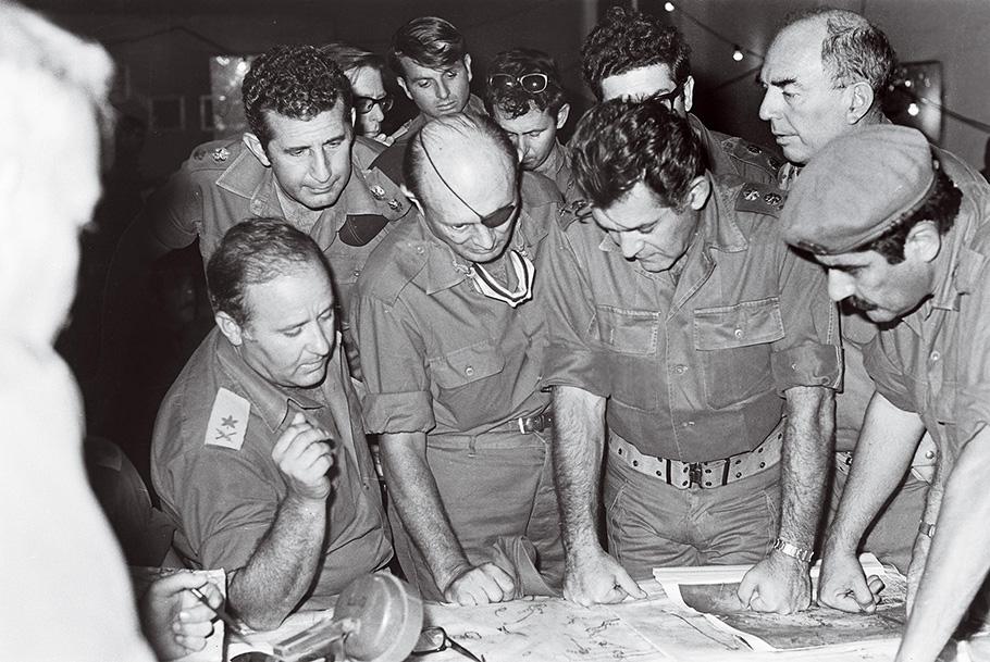 Dayan is surrounded by nine other officers, looking at a map on the table in front of them.