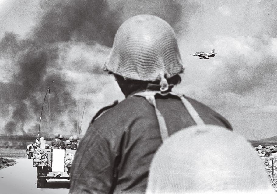 In this black and white photo, an aircraft can be seen streaking across a smoke-filled sky, while an Israeli soldier in the foreground looks on.
