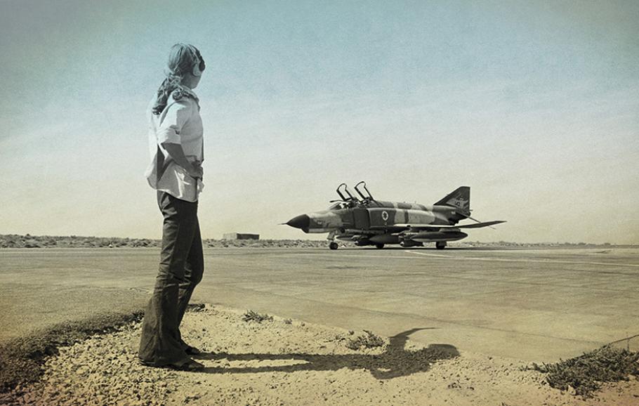 In this blue- and brown-tinged black and white photo, a member of the Israeli Air Force stands in the foreground of an airport, while a single aircraft sits on the runway.