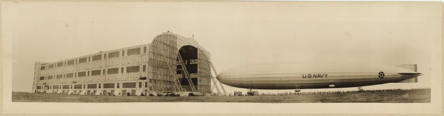  Panoramic photograph featuring a left side view of the US Navy airship Shenandoah under control of ground crew, being walked into its hangar.