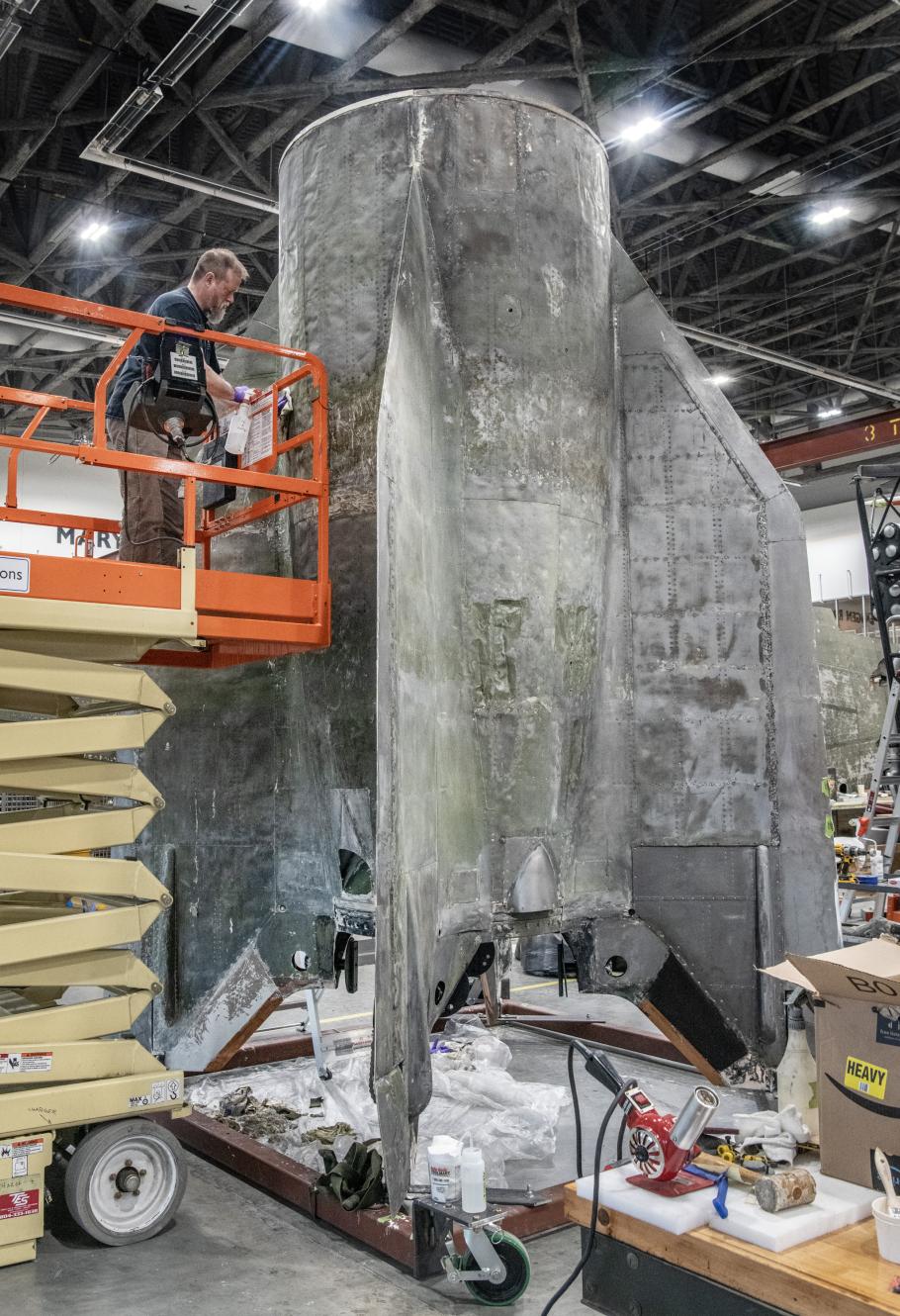 Large section of the bottom of a rocket is worked on inside a restoration facility.