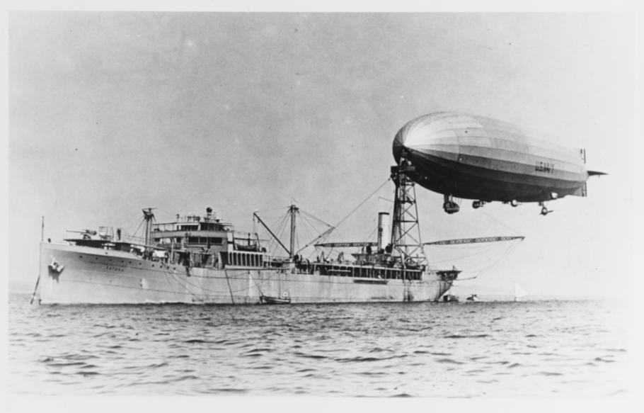 An image of a military ship at sea with a mooring mast. An airship is docked at the mast.