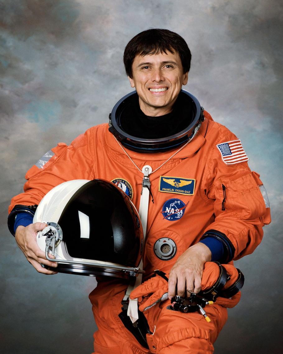 An astronaut holding a helmet in his hand and wearing a bright colored spacesuit smiles for a portrait.