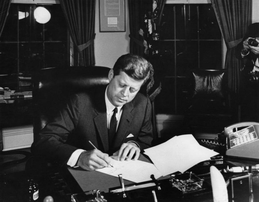 President Kennedy leans over a desk signing a document. 