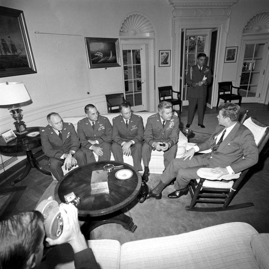 President Kennedy sits in a rocking chair, while four military pilots sit on a couch. 