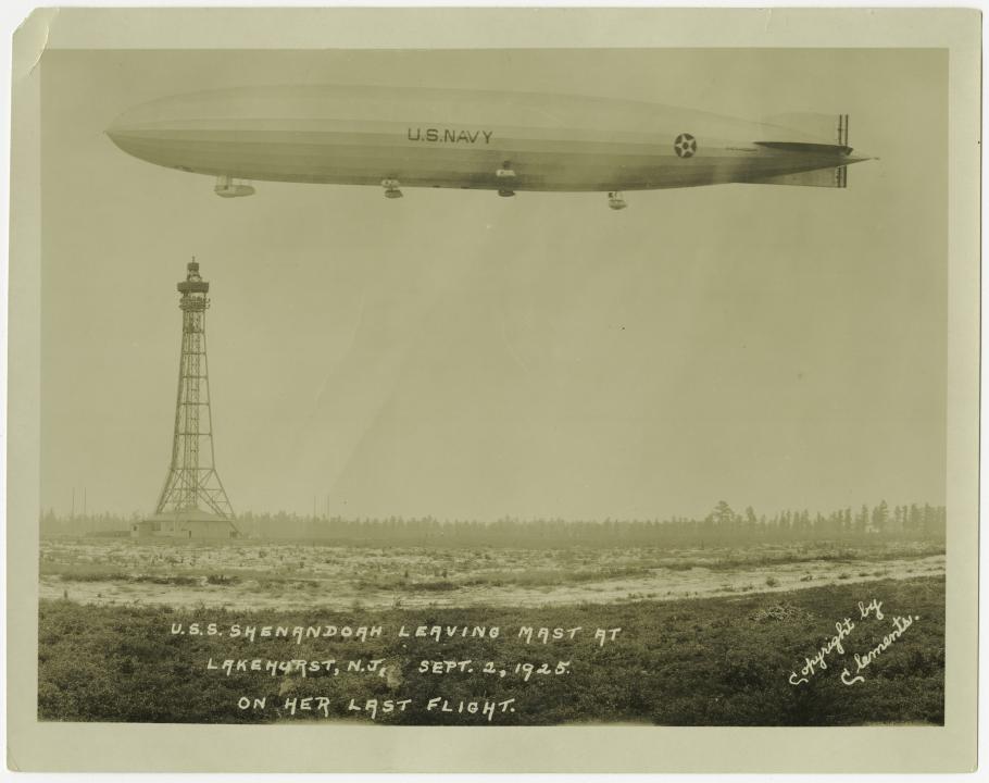 An airship floats in the air at the top of the frame. A mooring mast can be seen in the distance behind it.