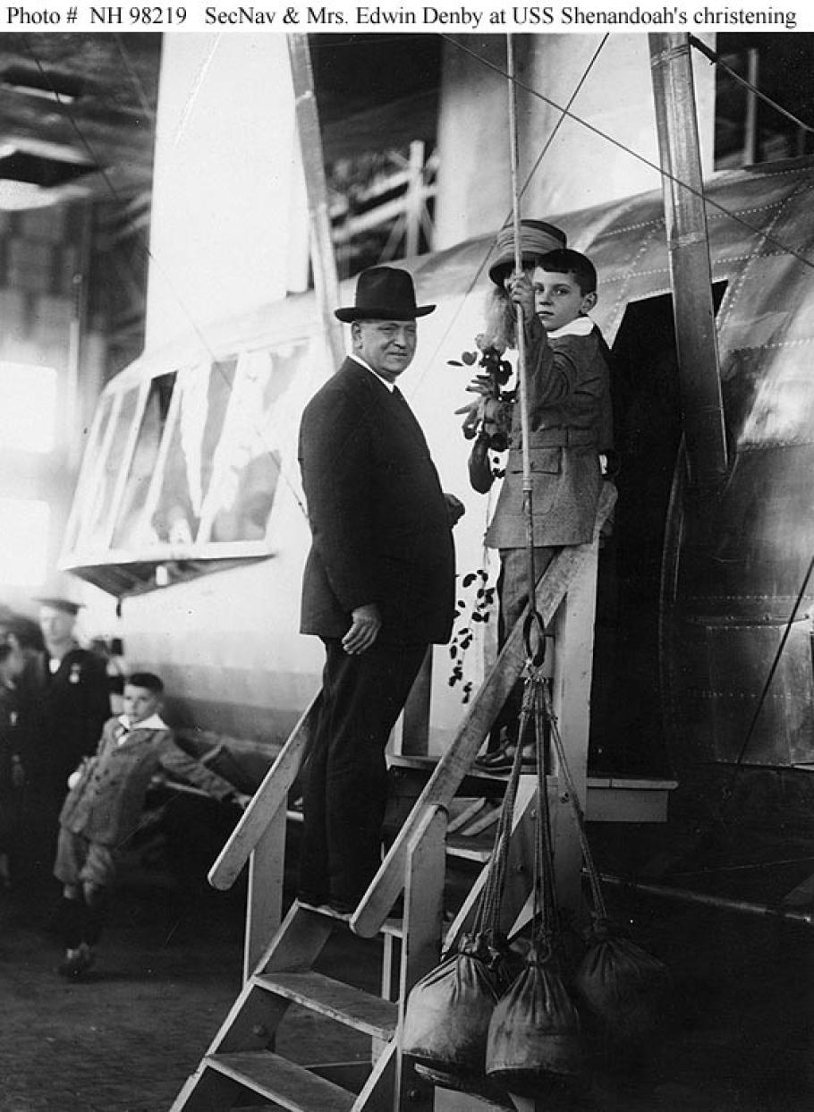 Three people stand on steps that lead to the entrance of an airship control car. They all look to the left, facing the camera.