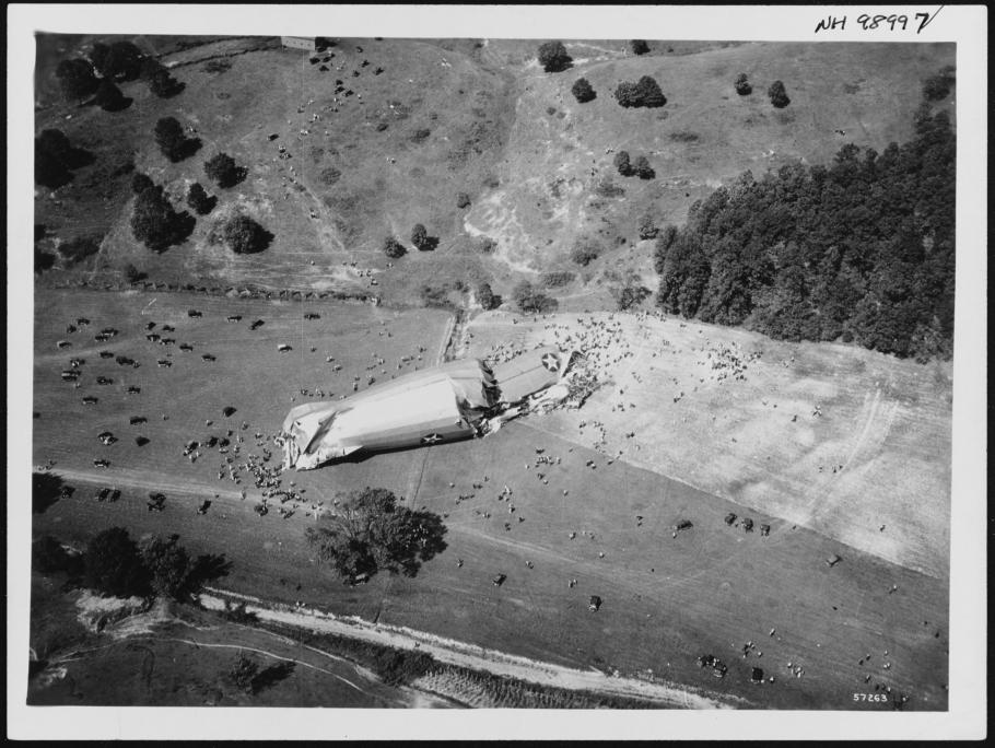 A cylindrical piece of an airship broken up in a field as seen from above. People crowed around it.
