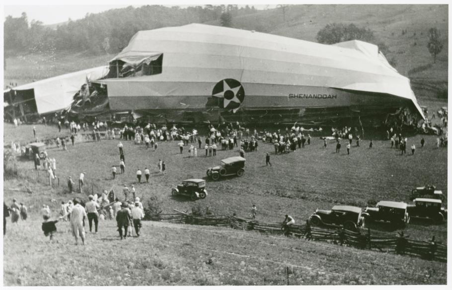 A wrecked piece of an airship sits in a fields as swarms of people flock to it.