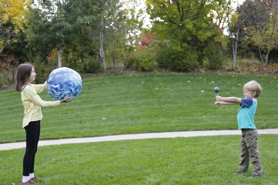 Two children stand on the grass about 6 feet away from each other. One holds a ball that represents the Earth. One holds a ball that represents the Moon. 