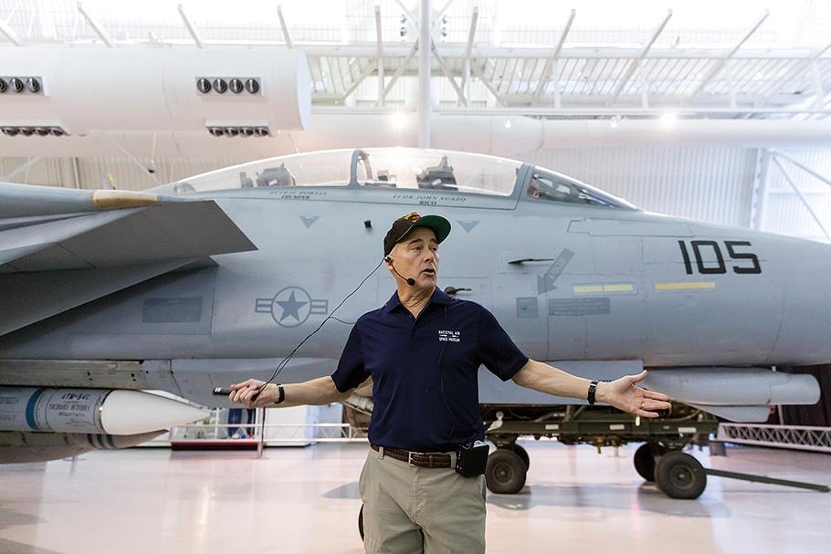 A middle-aged man in a polo shirt stands in front of a Grumman F-14, a large and sleek grey military airplane. Browne's arms are spread wide in a gesture, he wears a microphone, clearly engaging an audience.
