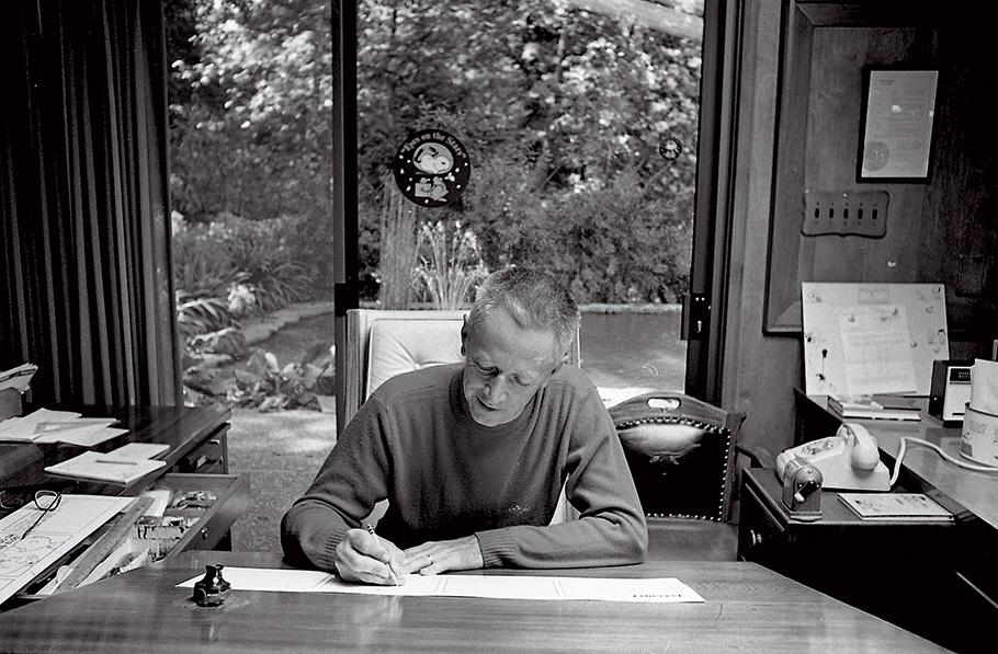 A black and white photograhp of cartoonist Charles Shulz, a middle-aged man in sweater, drawing a comic strip while sitting in a office in his California studio. In the background, glass doors offer a view into a yard, with a sticker depicting Snoopy, a cartoon beagle dog, as an astronaut stuck on the inside glass.