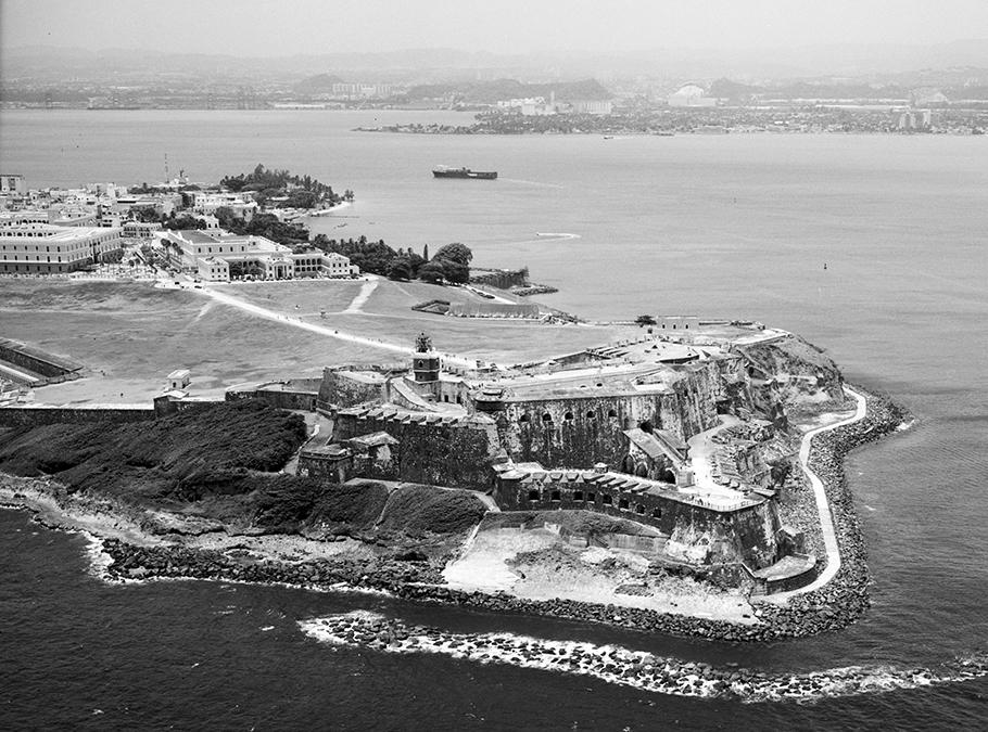 A black-and-white photo shows an aerial view of an island with a castle-like structure. 