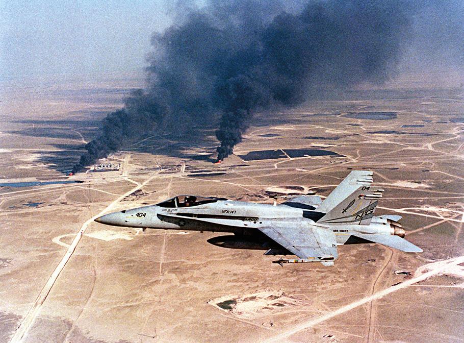 A silvery gray military jet armed with air-to-air missiles flys over a desert, as twin plumes of thick black smoke rise into the air.