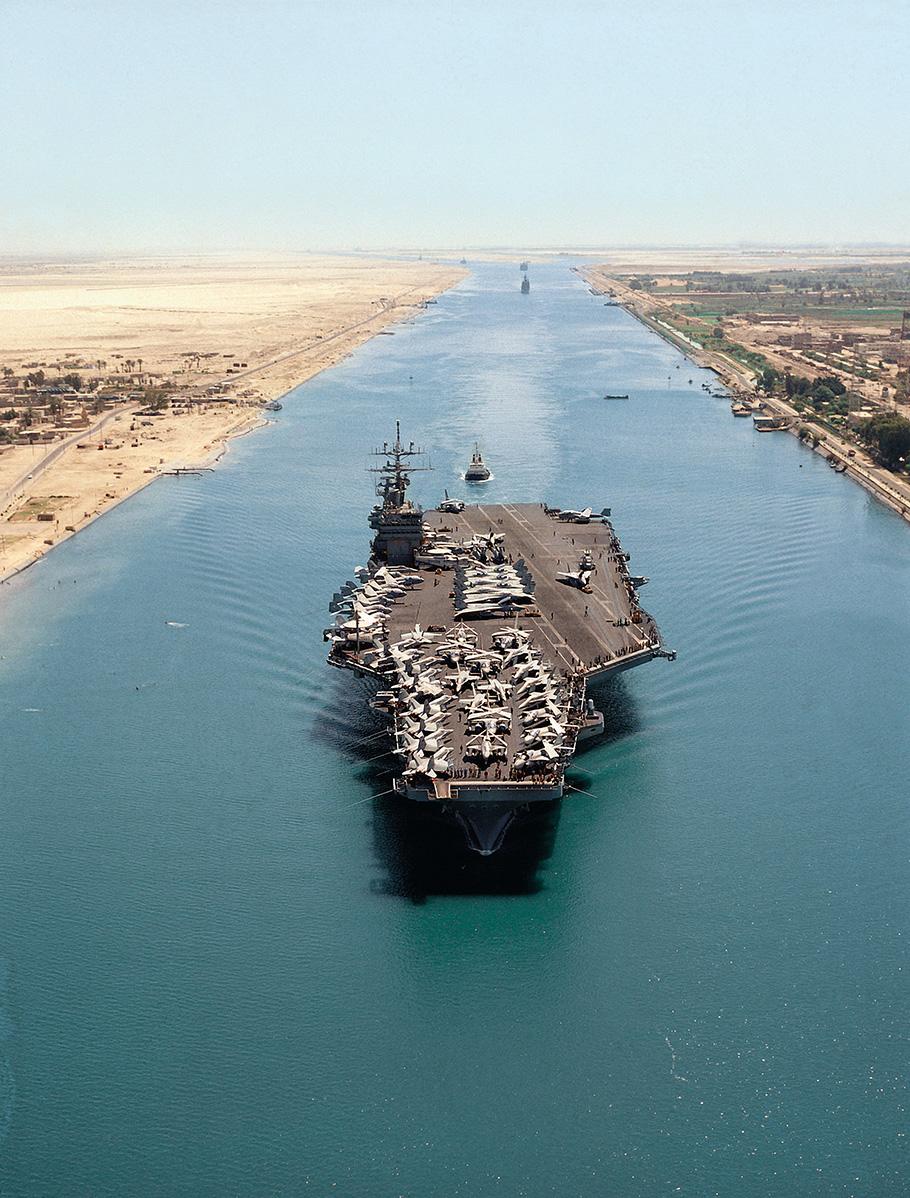A U.S. Navy aircraft carrier moves through a narrow body of water. Dozens of military aircraft are parked on the carrier's gray deck.