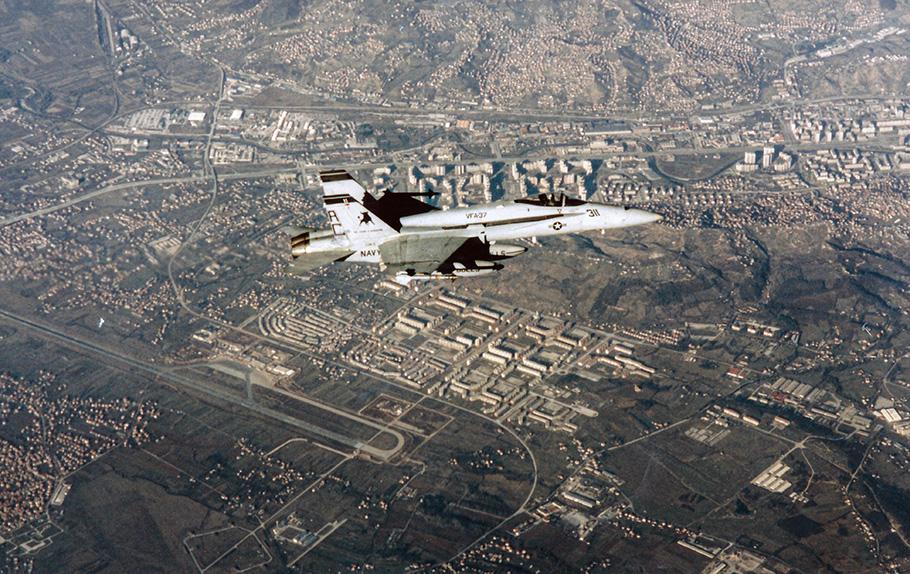 A light gray military jet armed with air-to-air missiles makes a low-altitude pass over a city.