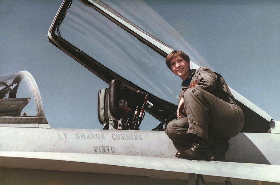 A young woman wearing a U.S. Navy olive-green flightsuit and black combat boots perches next to the cockpit of her F/A-18C miltary jet. "Lt Sharon Cummins" is stenciled onto the side of the airplane.