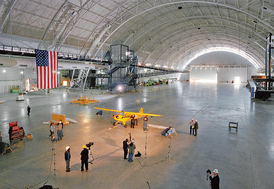 A bright yellow Pipe J-3 Cub sits alone in a vast empty hangar with an American flag hanging from the ceiling. 