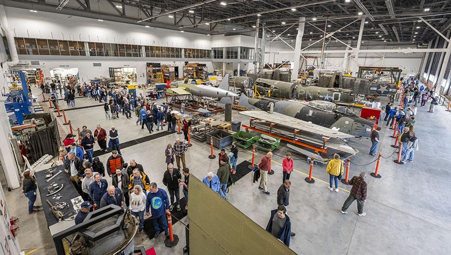 A crowd of people explore a hangar with concrete floors and aircraft in various stages of completion and restoration. There are windows at the top of the hangar allowing people on the second floor to peer inside.