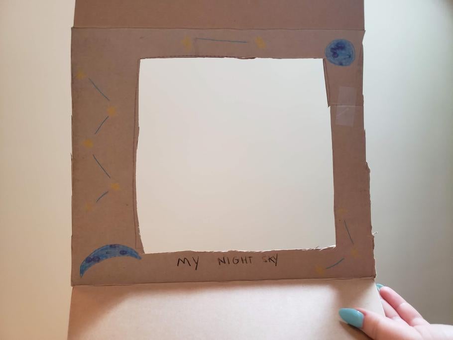  Square piece of cardboard with a square opening cut into the middle of the cardboard. A planet, a moon, and yellow and blue decorations have been hand drawn around the border of the opening. The words, "My Night Sky" are hand written on the bottom of the square opening. 