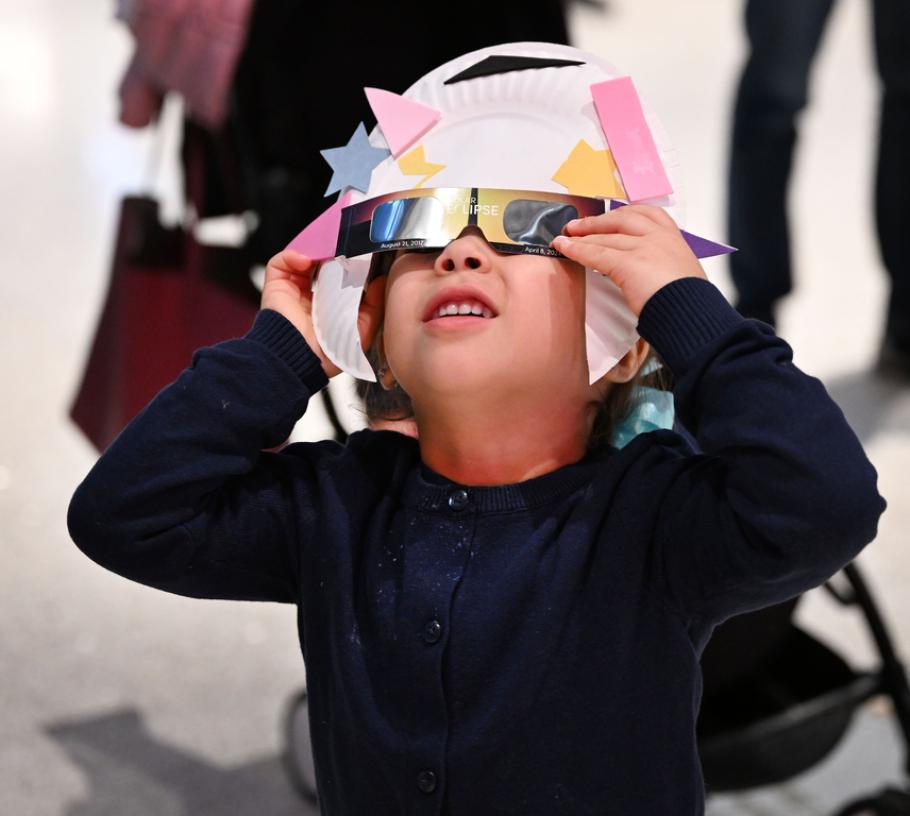 A young visitor uses an eclipse viewer they made from eclipse glasses and a paper plate to safely view an annular eclipse. They squint up in concentration. 