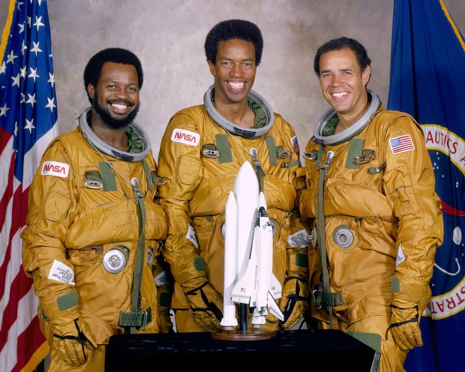 Three Black men wearing yellow-brown astronaut flight suits. A model of the space shuttle is in front of them.