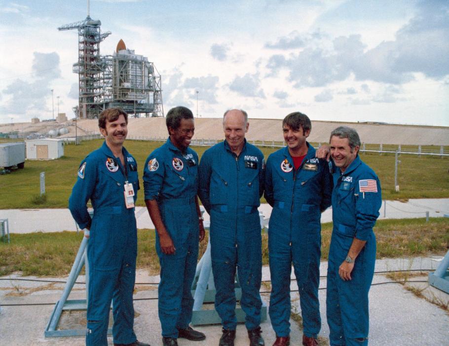 Five men in blue NASA flight suits stand grouped together. A launch pad with a space shuttle stack on it is seen in the distance behind them.