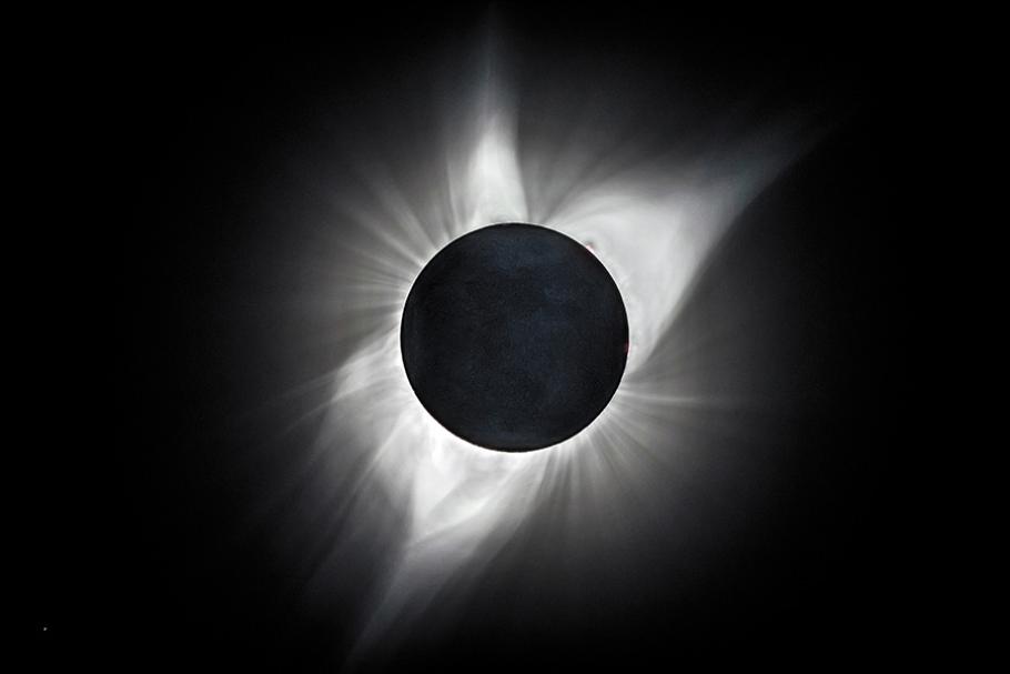 A black-and-white image of a total solar eclipse shows the sun's bright corona peaking out from the edges of the moon.