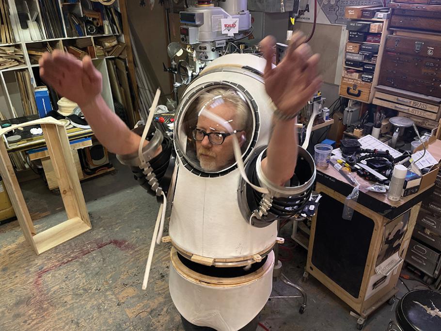Adam Savage tries on the white fiberglass upper torso of his spacesuit, which is fitted with sleeves made from black plastic bands. His arms are held above his head as he tests the suit's mobility.