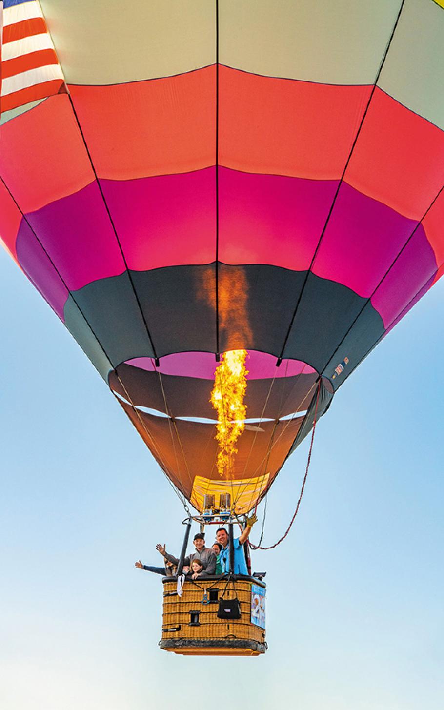 In mid-air, a black-, magenta-, and coral-colored balloon is inflated by a long flame from the burner. Suspended below the balloon is a wicker basket carrying at least five people, men and women and one girl. The people are waving and smiling. 