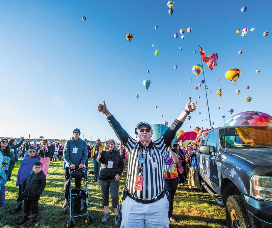 A middle-age woman wearing khaki pants and a black-and-white stripe shirt stands on the ground--below dozens of hot air balloons flying above. Both of her arms are raised.