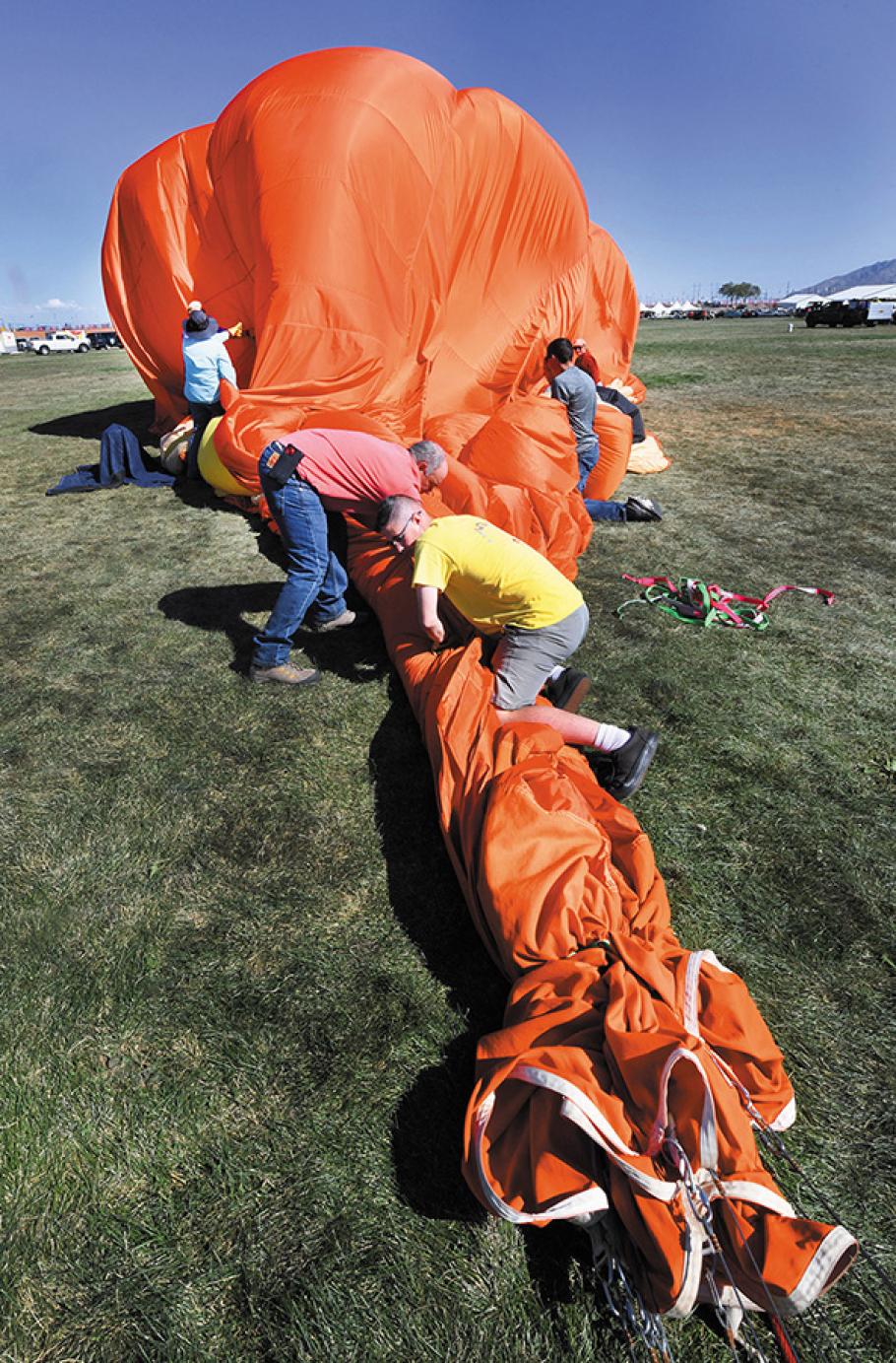 The partially-deflated envelope of a brightly-colored balloon is stretched out on the grass as four people work to squeeze out the remaining air, under the direction of middle-age white man standing off to the side.