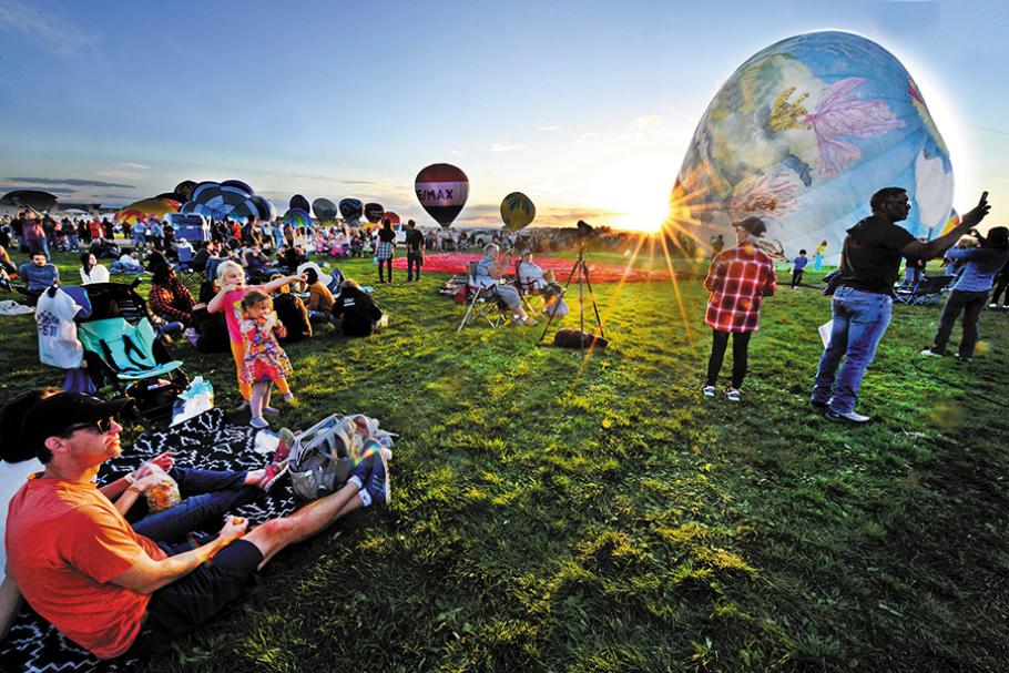 With the sun beaming low on the horizon, dozens of people are sitting or standing on an expanse of grass, framed by several balloons waiting to take off into the sky.